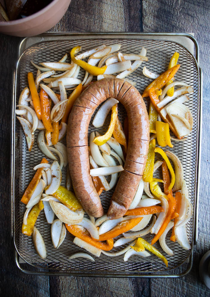 raw hasselback kielbasa on an air fryer basket surrounded by raw onions and peppers