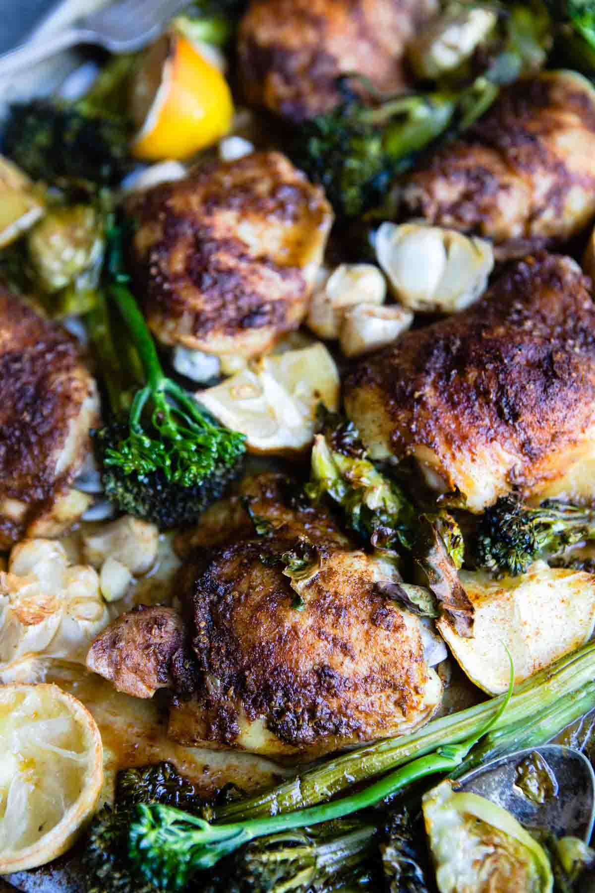 close up of baked chicken thighs with a paprika crust next to roasted veggies and lemons and garlic