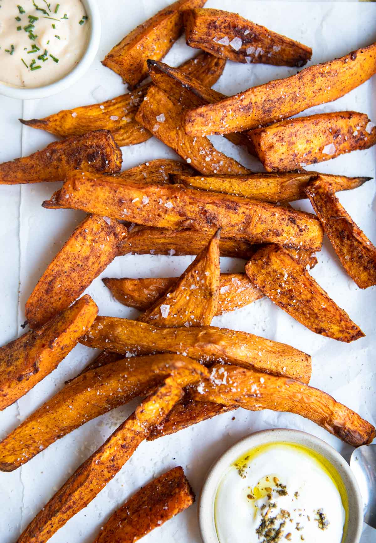 crispy air fryer sweet potato wedges sprinkled with sea salt and sitting on a piece of parchment paper next to dipping sauces 