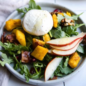 gray plate filled with roasted pumpkin salad and topped with bacon and burrata cheese, pear and pomegranate seeds