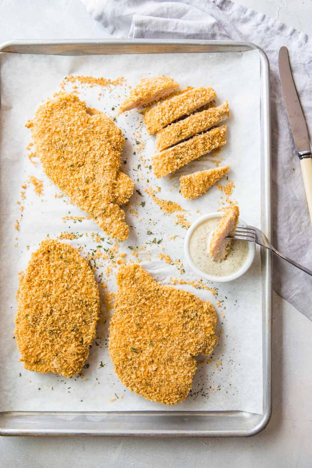 baked chicken cutlets on a baking sheet with one cutlet sliced and being dipped in ranch dressing 