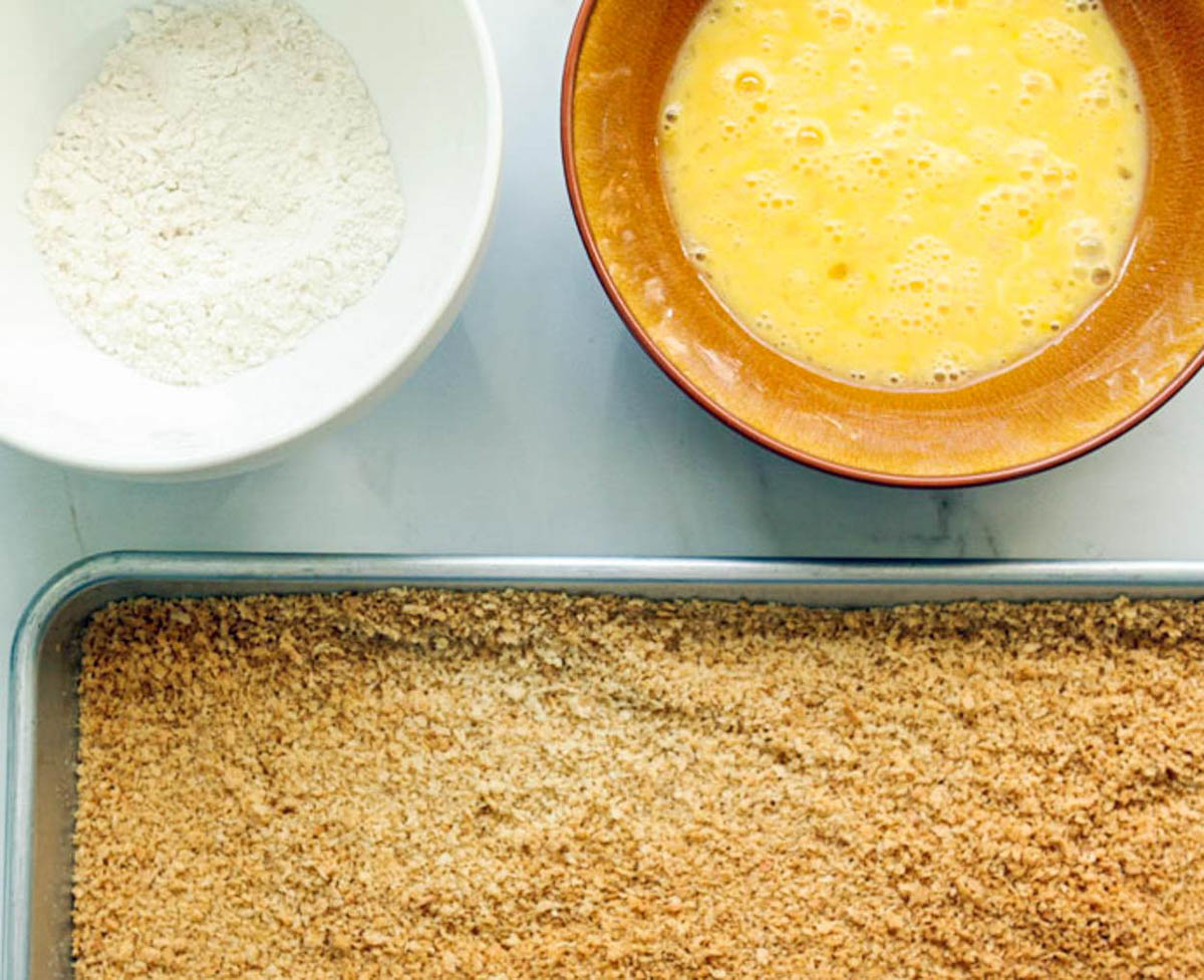 2 shallow bowls one filled with flour the other filled with whisked eggs, next to a baking sheet filled with breadcrumbs 