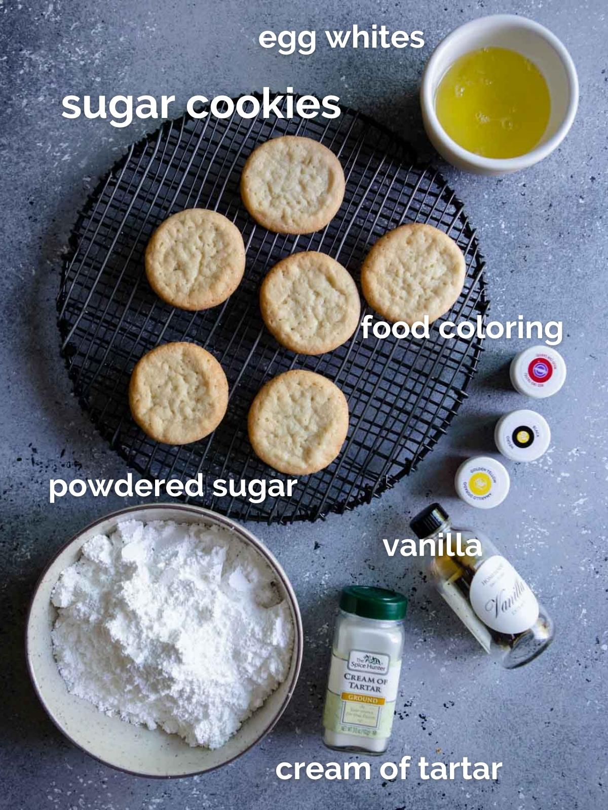 baked sugar cookies, a bowl of powdered sugar, a bowl of egg whites, a jar of cream of tartar, vanilla extract and food colorings 