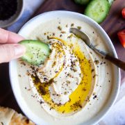 a woman dipping a cucumber in a bowl of high protein hummus swirled with olive oil