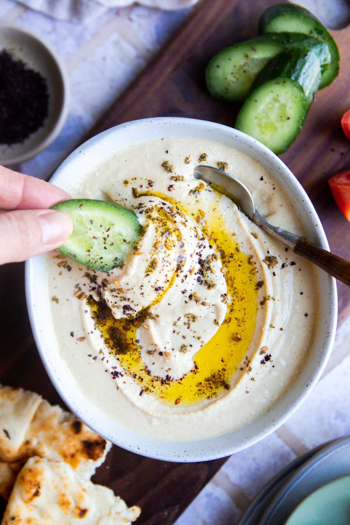 a woman dipping a cucumber in a bowl of high protein hummus swirled with olive oil 