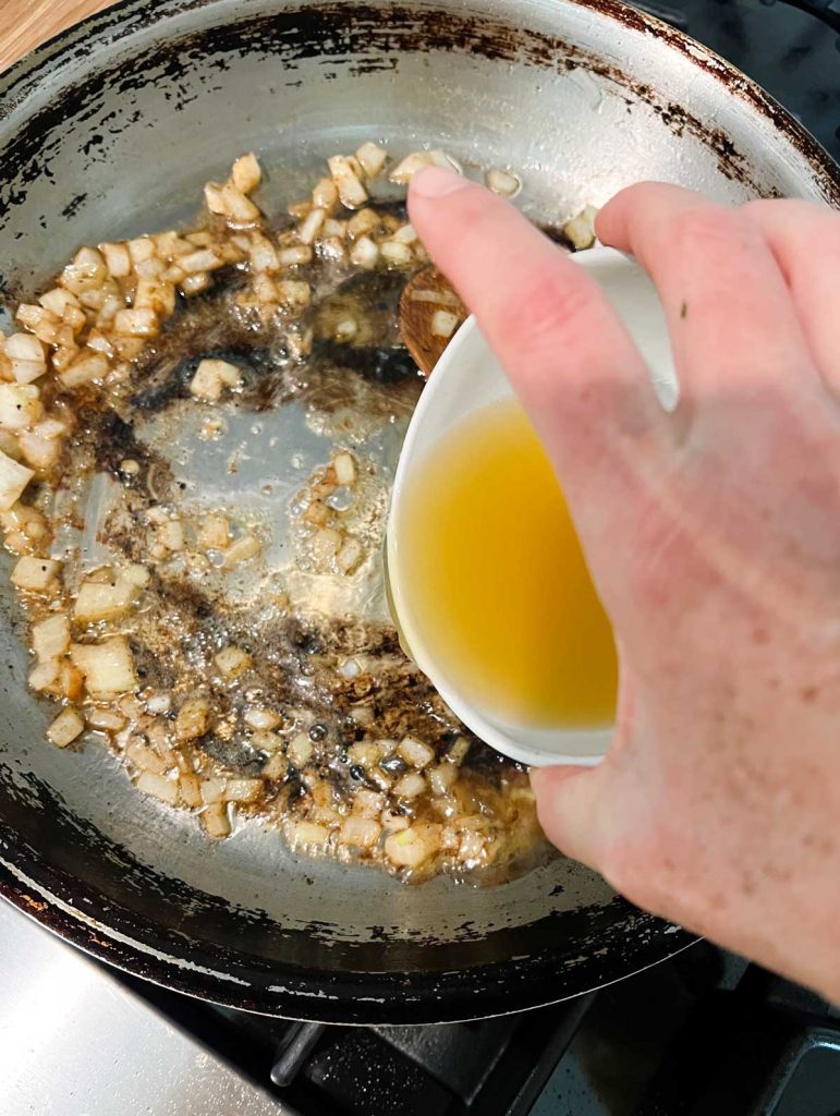 chicken broth being poured into a skillet with sauteed onions 