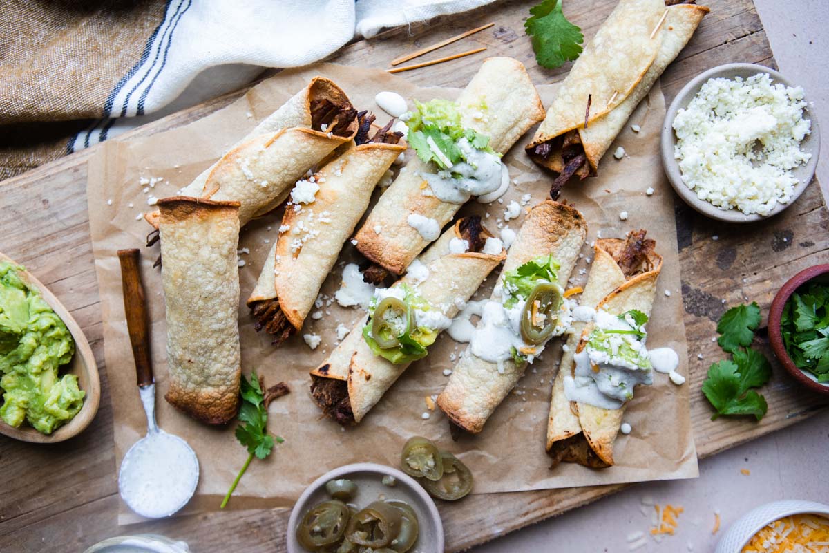cutting board with parchment paper set on top and beef taquitos garnished with various toppings on the parchment paper
