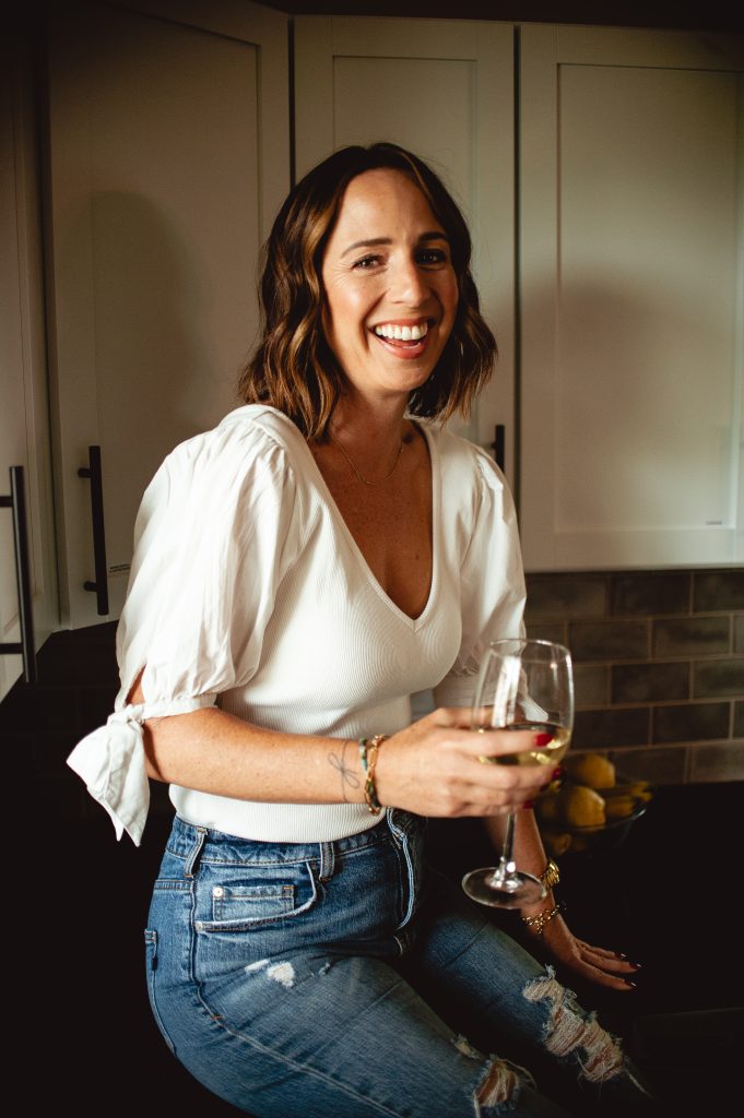 Caucasian women sitting on a kitchen counter smiling, holding a glass of white wine 