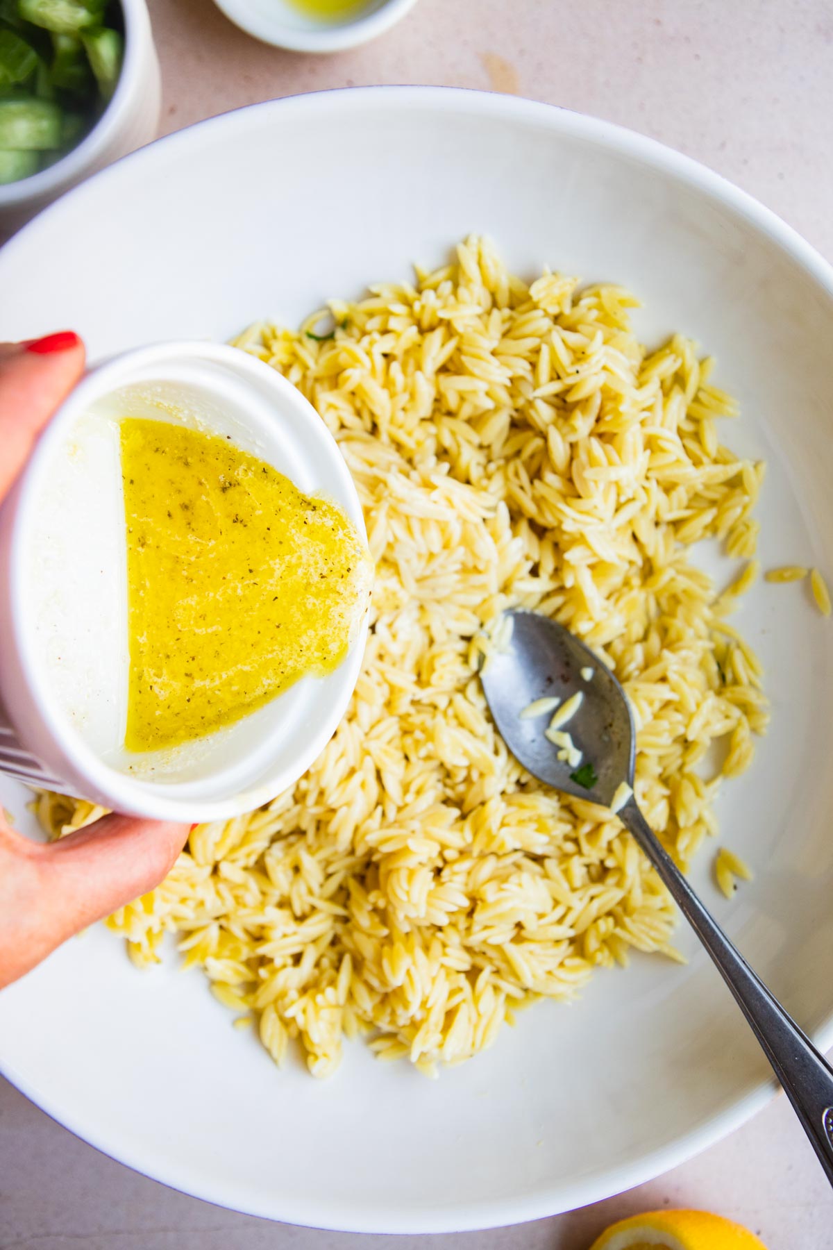 Hand pouring lemon parmesan dressing over a bowl of cooked orzo pasta.