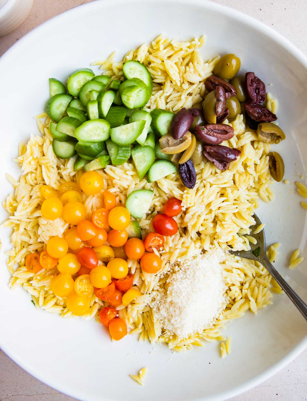 Chopped veggies and olives halved in a bowl with cooked orzo and parmesan cheese before combining.