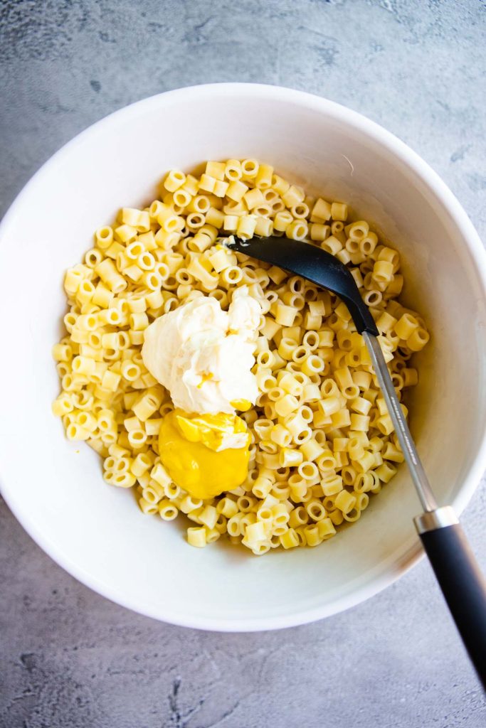 Mayo and mustard measured out over a bowl of cooked ditalini pasta in a large bowl.