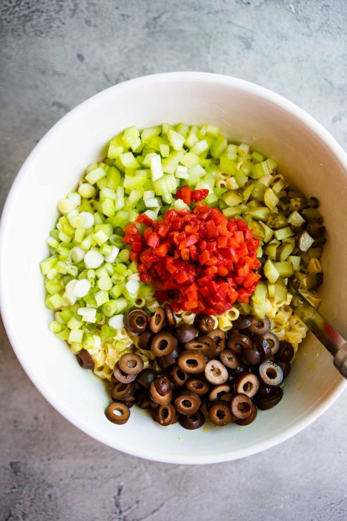 Chopped veggies and black olives added over cooked ditalini pasta in a large mixing bowl.