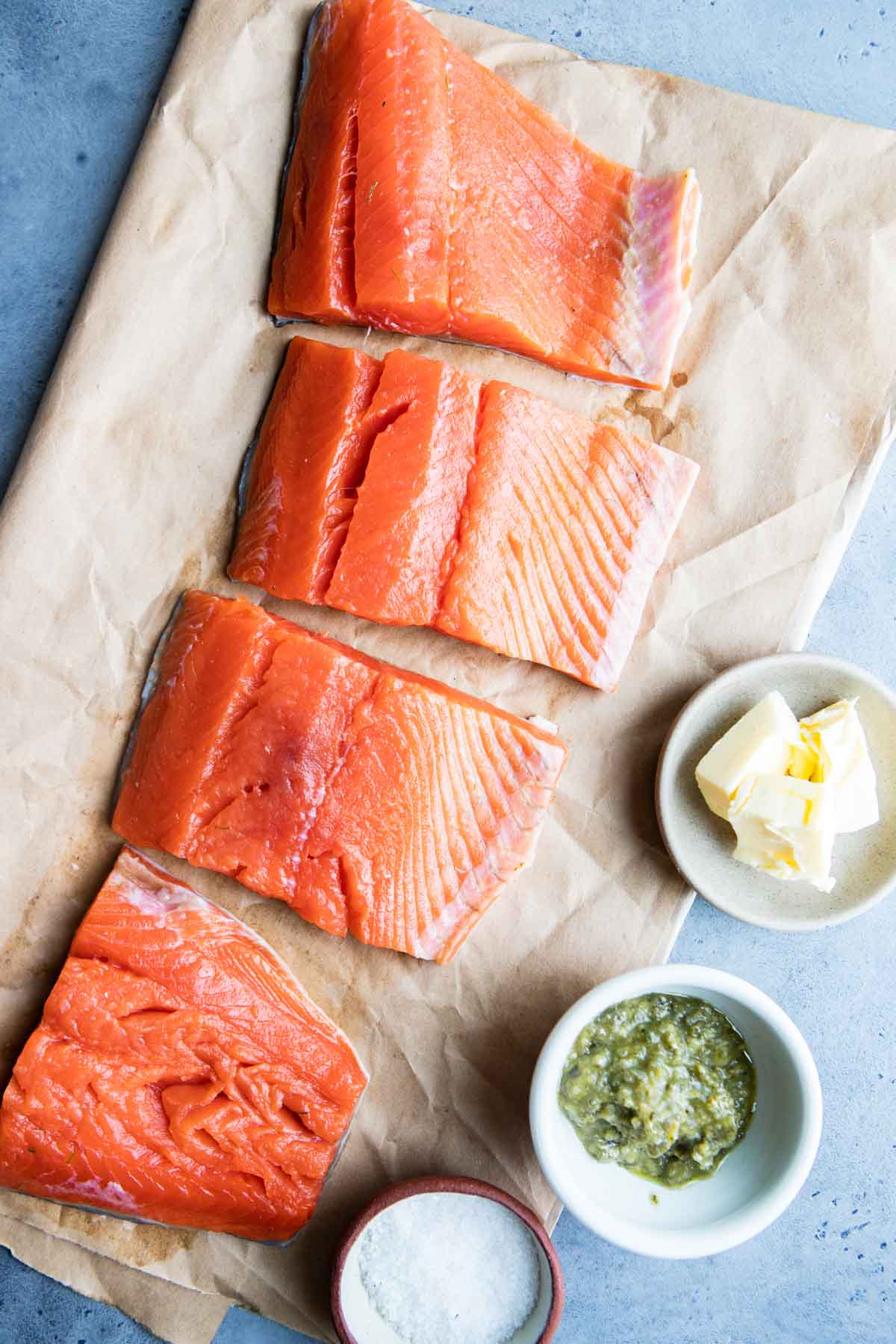 Salmon on parchment paper and the remaining ingredients for pesto butter salmon measured into small bowls.