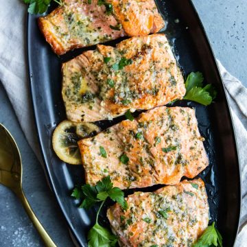 Pesto butter salmon garnished with parsley, served on a serving platter with lemon slices and a serving utensil resting on the side.