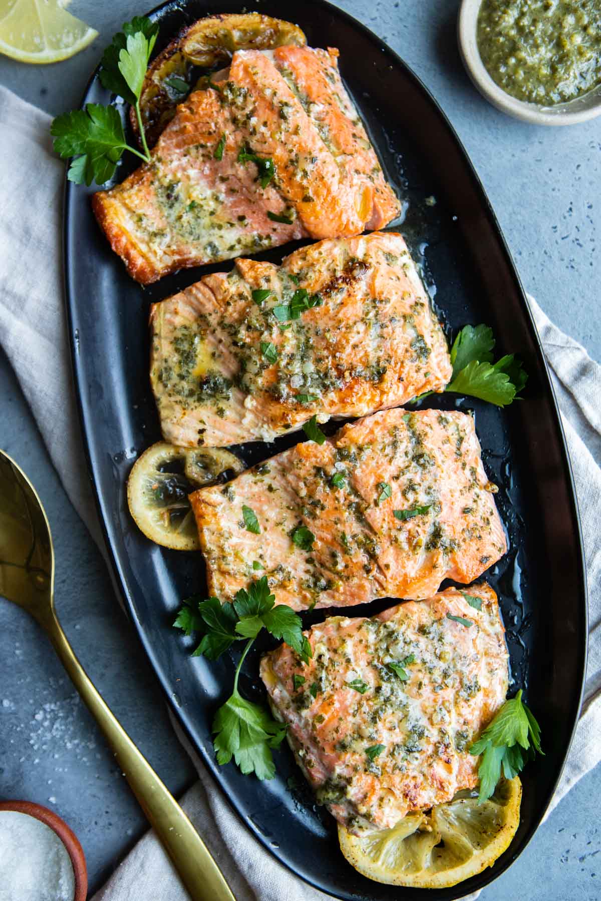Pesto butter salmon garnished with parsley, served on a serving platter with lemon slices and a serving utensil resting on the side.