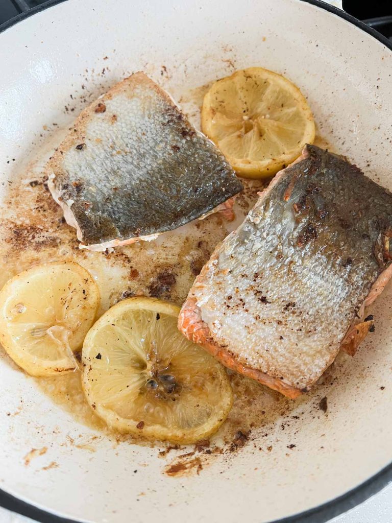 Lemon slices laying next to salmon filets skin side up in a frying pan.