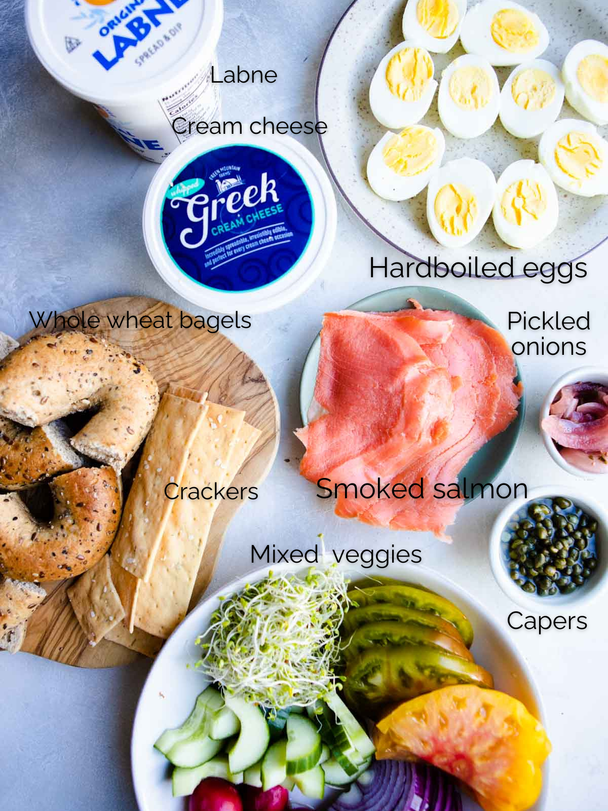 smoked salmon, hardboiled eggs, sliced tomatoes and onions, cream cheese and capers in small bowls, ready to make a grazing board