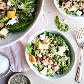 a small bowl filled with arugula quinoa salad topped with apples, goat cheese and hazelnuts