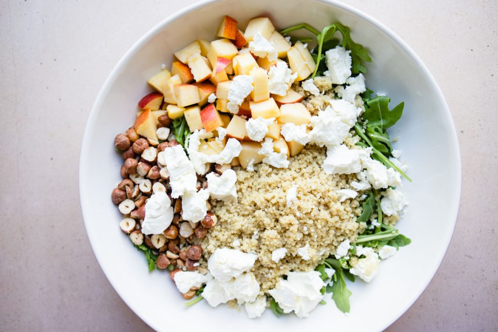 chopped apples, cooked quinoa, arugula, toasted hazelnuts and arugula in a large white bowl