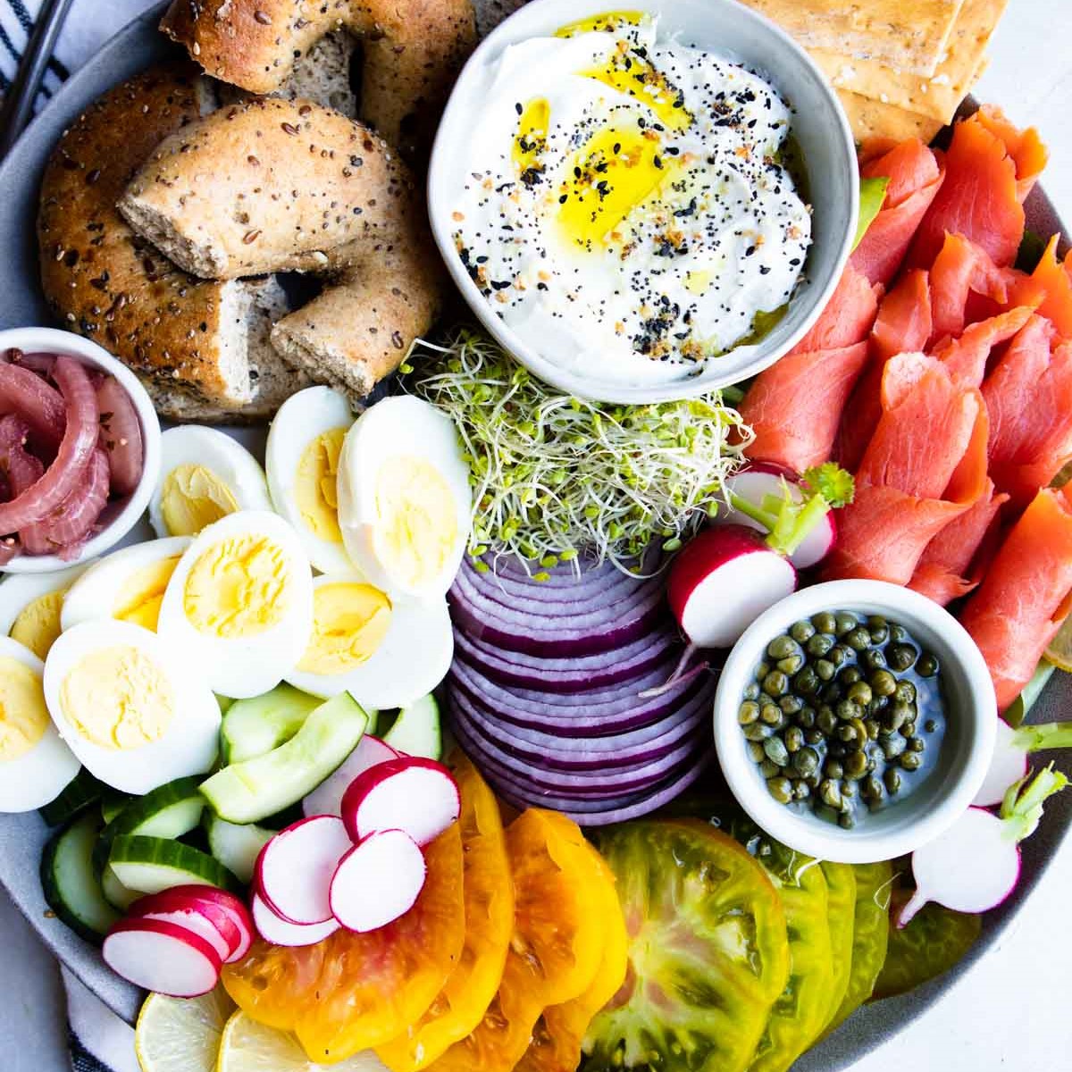 smoked salmon rolled up on a round platter along with sliced veggies, capers, cut up bagels and yogurt sauce