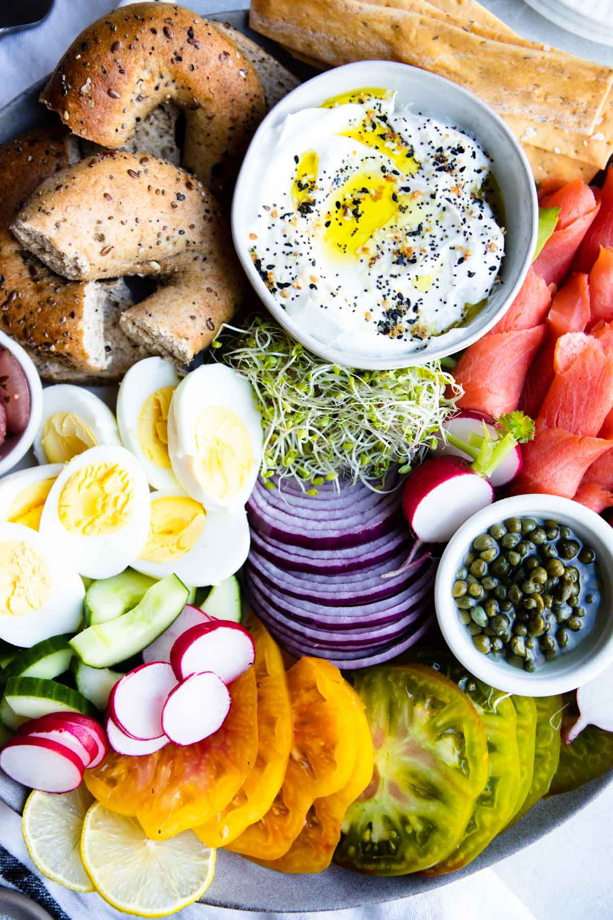 labneh, bagels, hardboiled eggs, sliced tomatoes and red onions and cucumbers and smoked salmon on a gray platter 