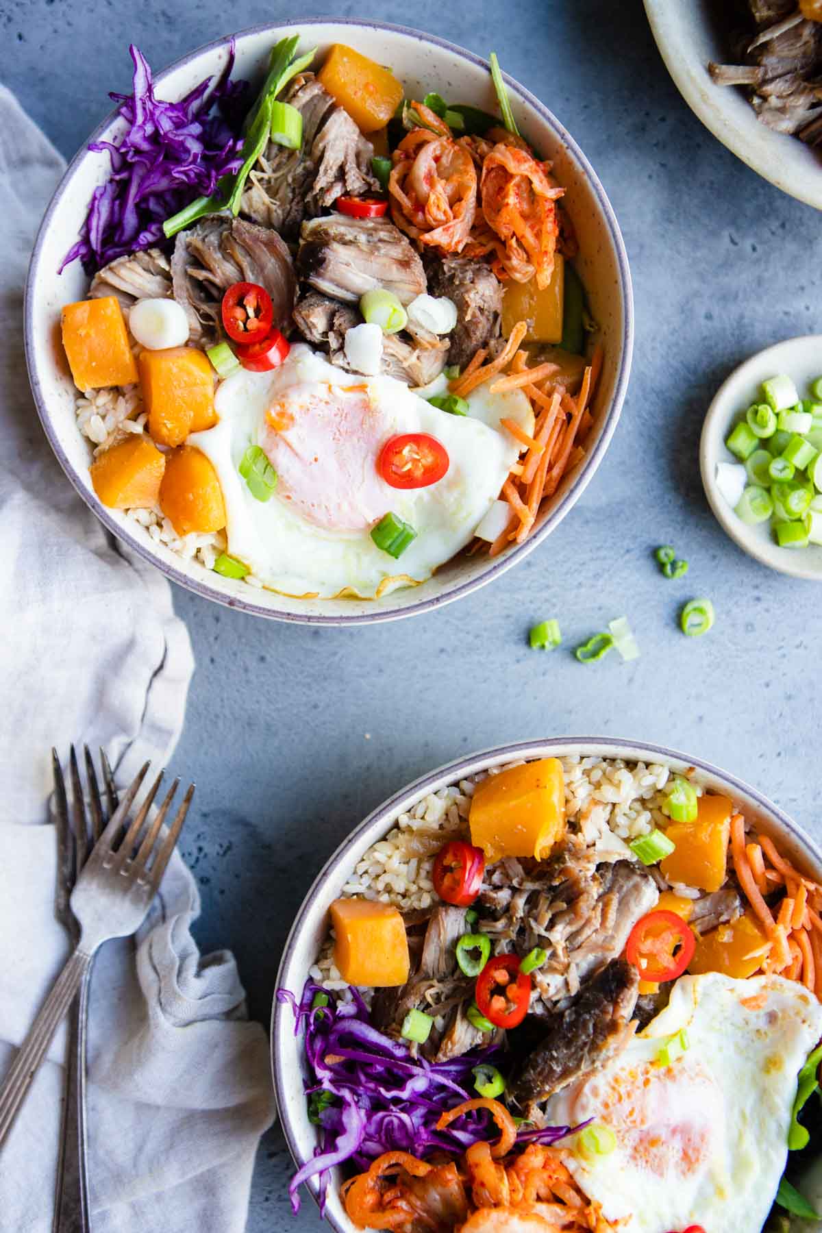 2 bowls filled with shredded pork and squash topped with veggie garnishes, kimchi and an over easy egg 