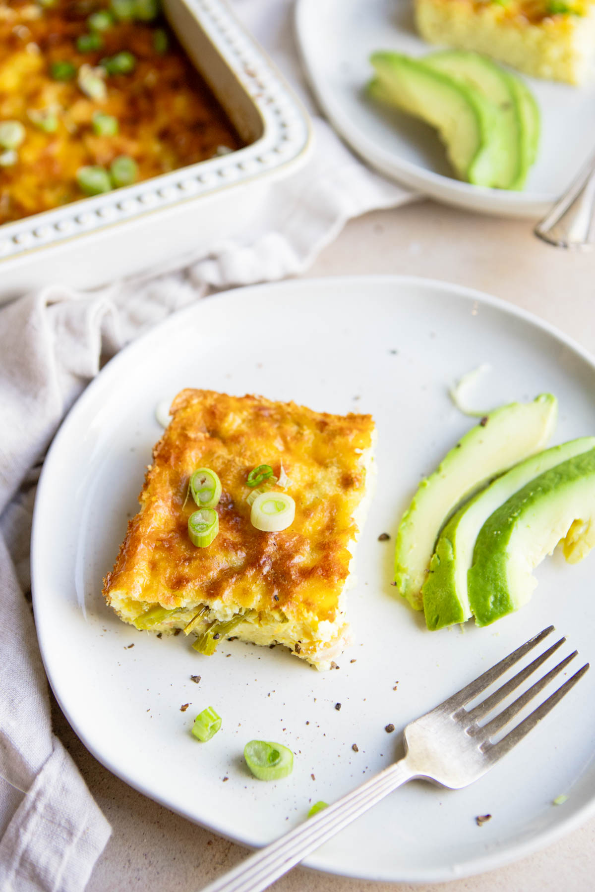 one piece of baked eggs and asparagus casserole on a white plate next to 3 slices of avocado 