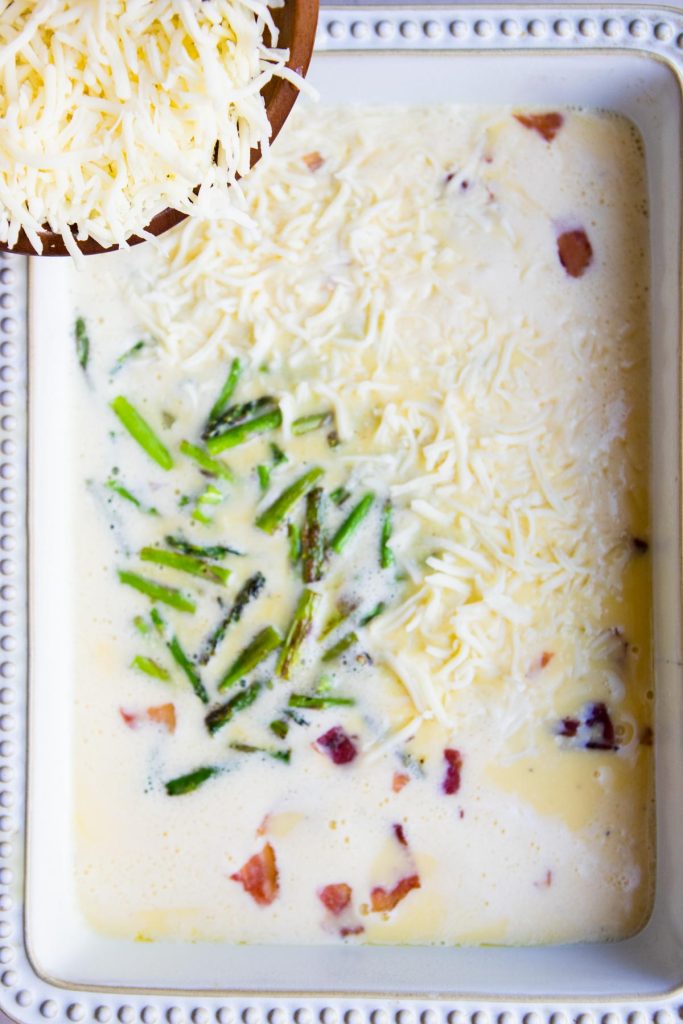 blended eggs poured in a casserole dish with asparagus and bacon and topped with shredded cheese 