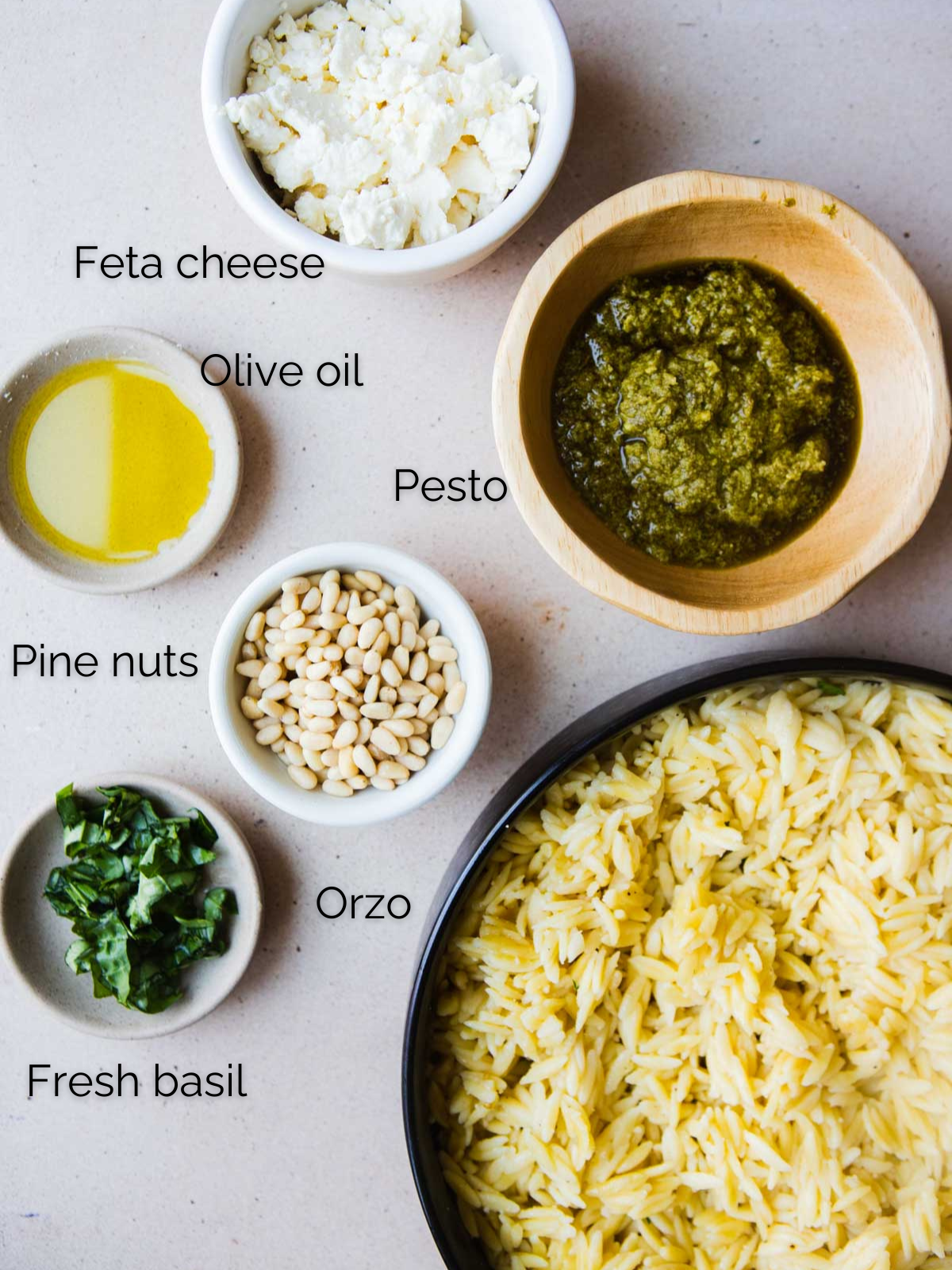 cooked orzo pasta in a black bowl, feta cheese, pesto sauce, pine nuts, olive oil and basil all in small bowls