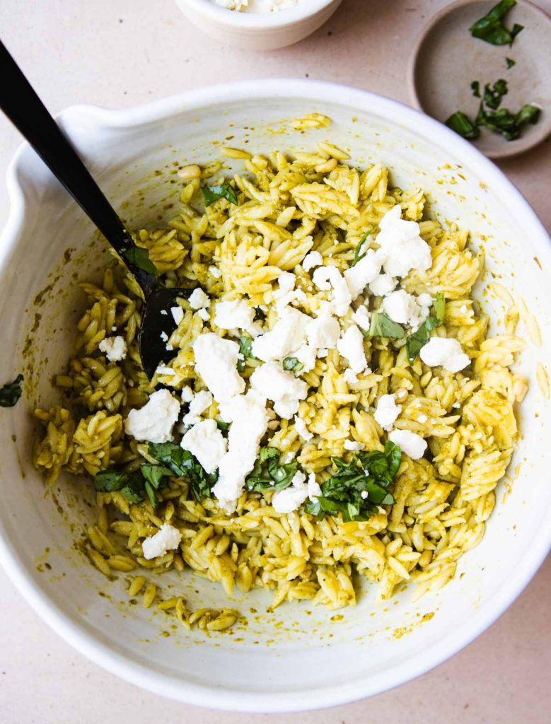 feta cheese and fresh basil being added to a pesto orzo salad