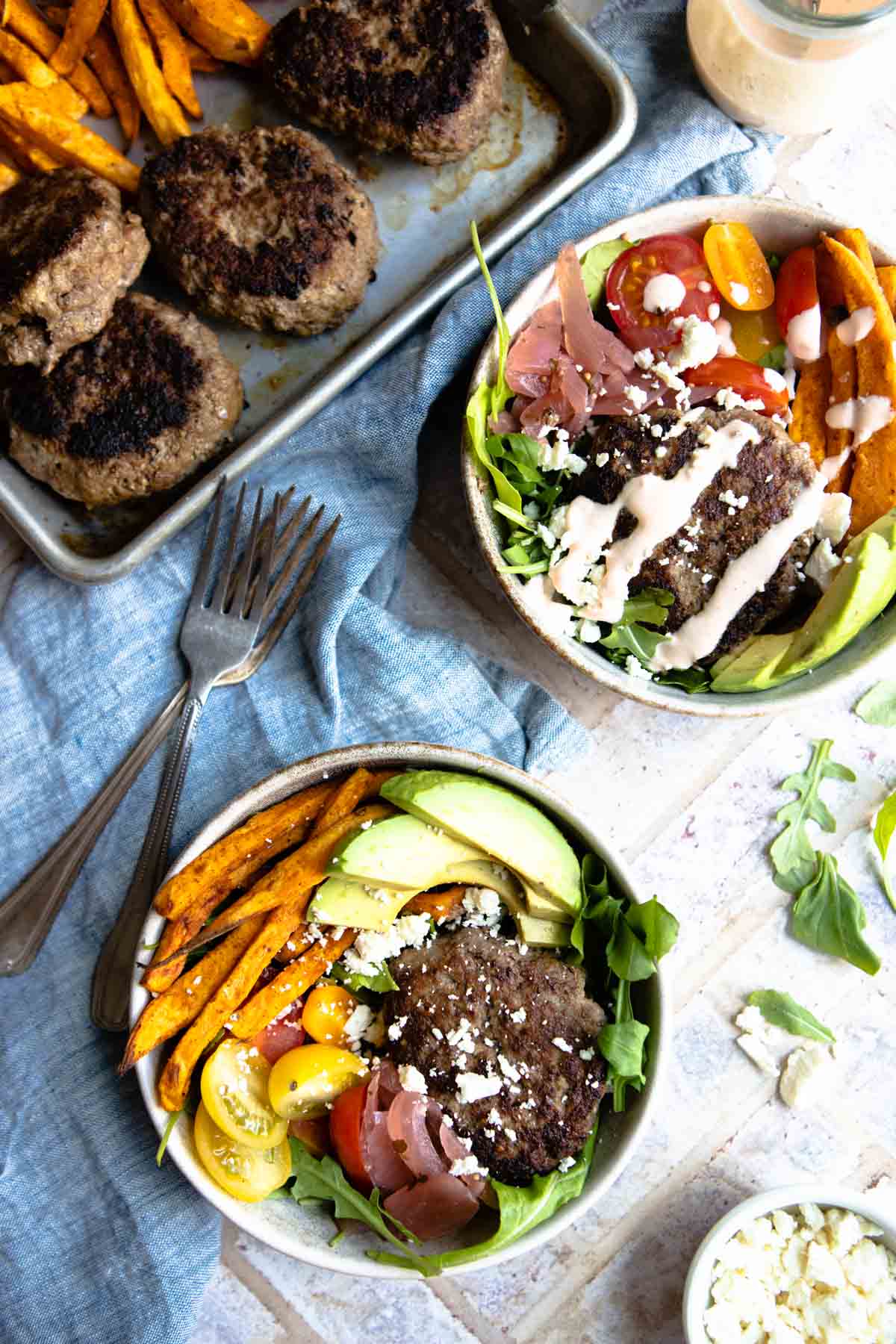 hamburger bowls in beige bowls next to a sheet pan with cooked burgers and sweet potato wedges