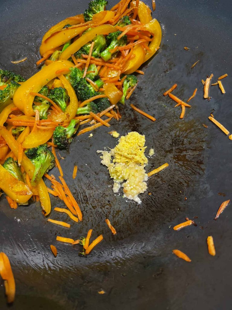 carrots and other veggies being cooked in a wok, with ginger and garlic added in