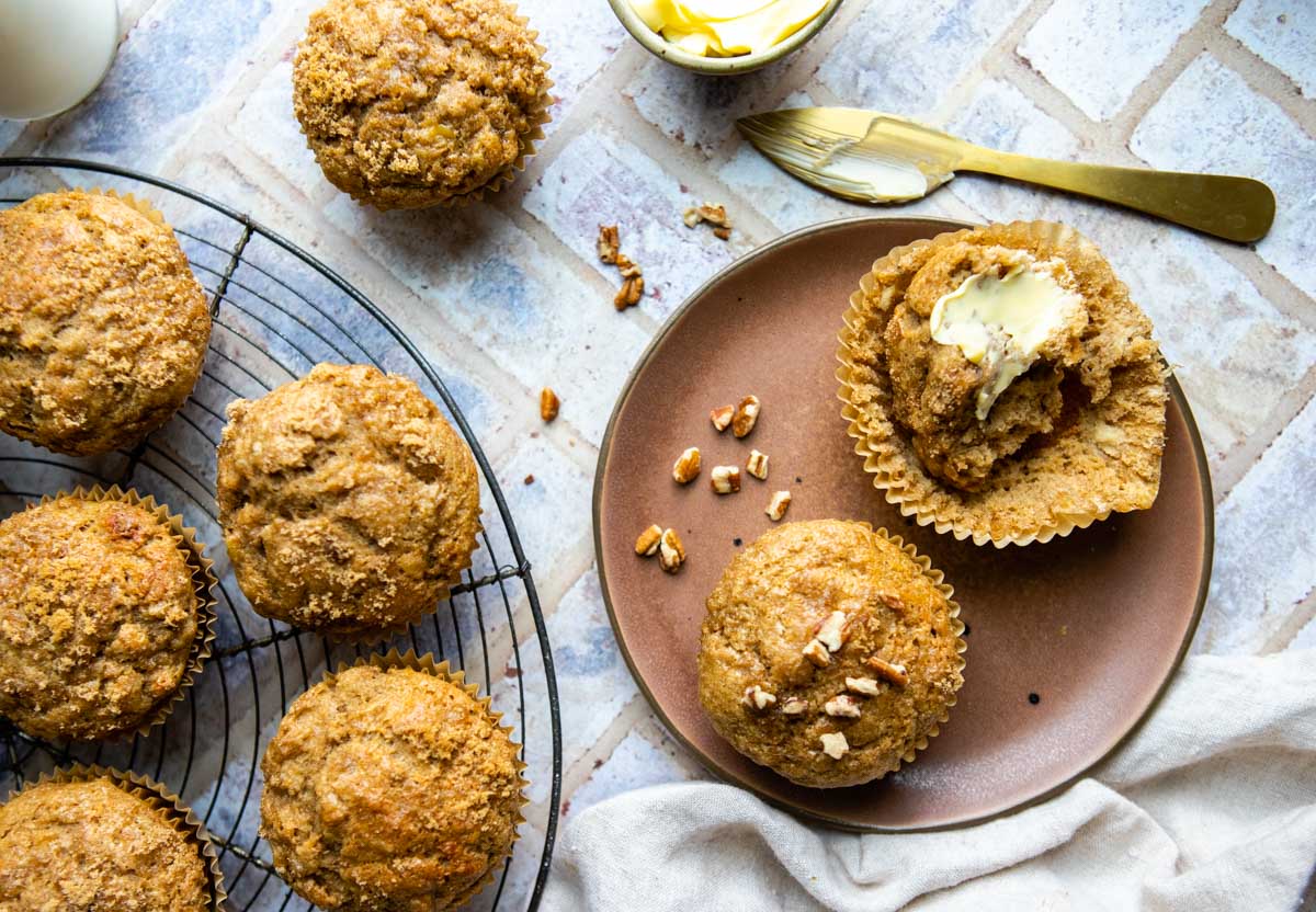 round wire cooling rack with banana protein muffins and 2 muffins on a brown plate with 1 muffin spread with butter