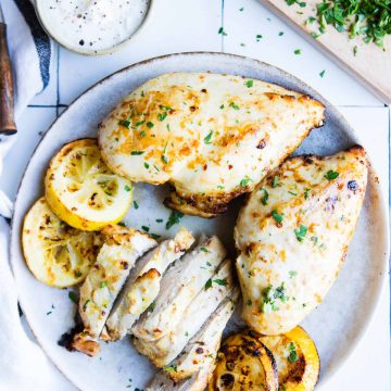 whole and sliced air fryer lemon chicken breasts on a gray plate garnished with grilled lemons