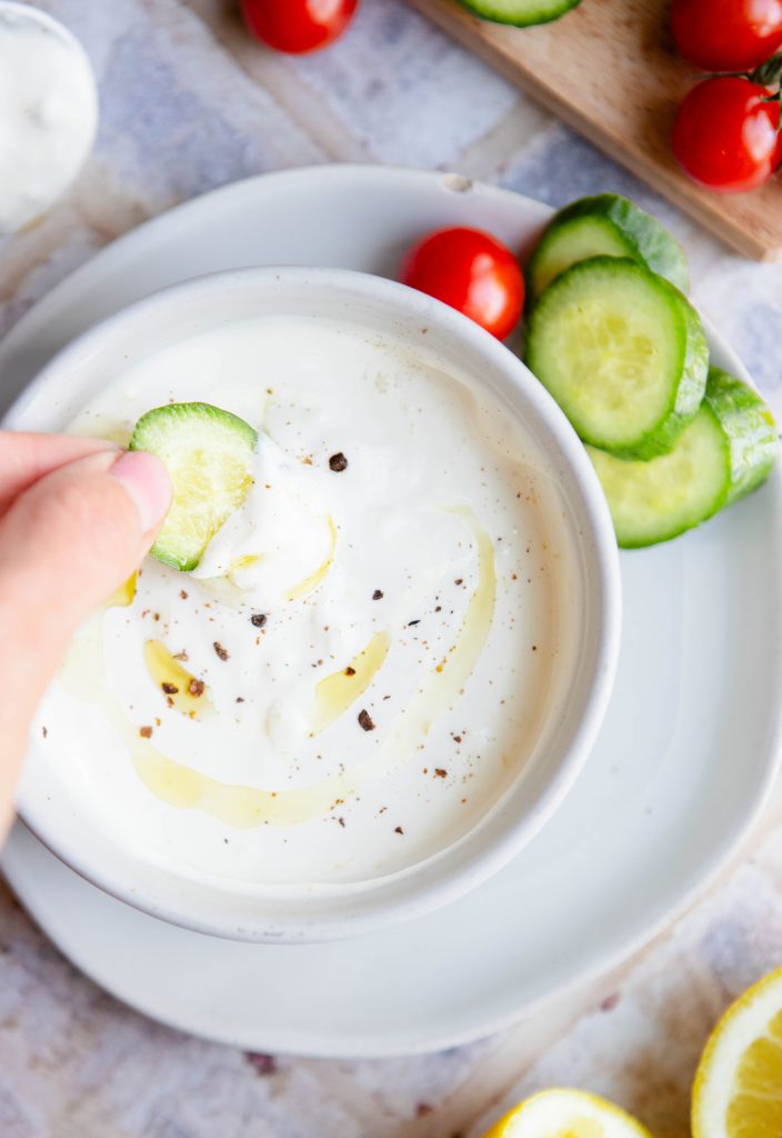 garlic yogurt sauce drizzled with olive oil and black pepper in a white bowl and a women's hand dipping fresh cucumbers into it