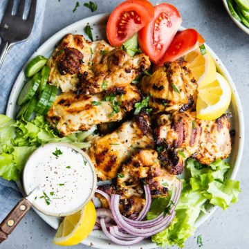 grilled chicken thighs on a plate next to a bowl of garlic yogurt sauce and other veggie garnishes