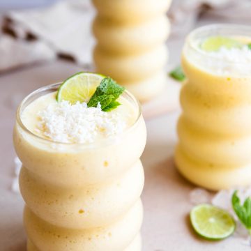 3 wavy glasses filled with a mango protein smoothie and garnished with lime and shredded coconut