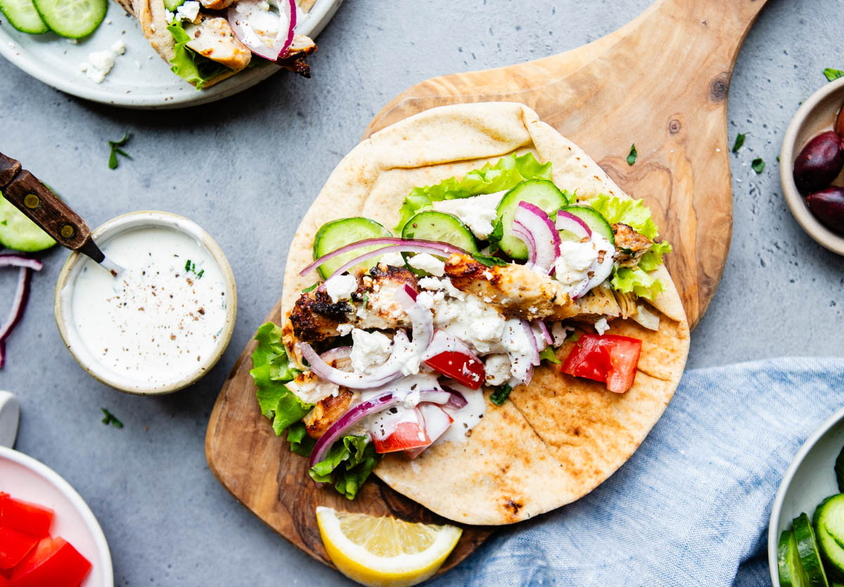 a wood board with a chicken wrap on top and garnished with cucumbers olives and garlic yogurt sauce