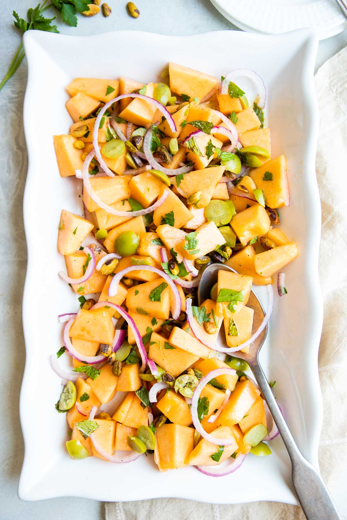 melon salad tossed with green olives and red onions on a white ceramic serving platter