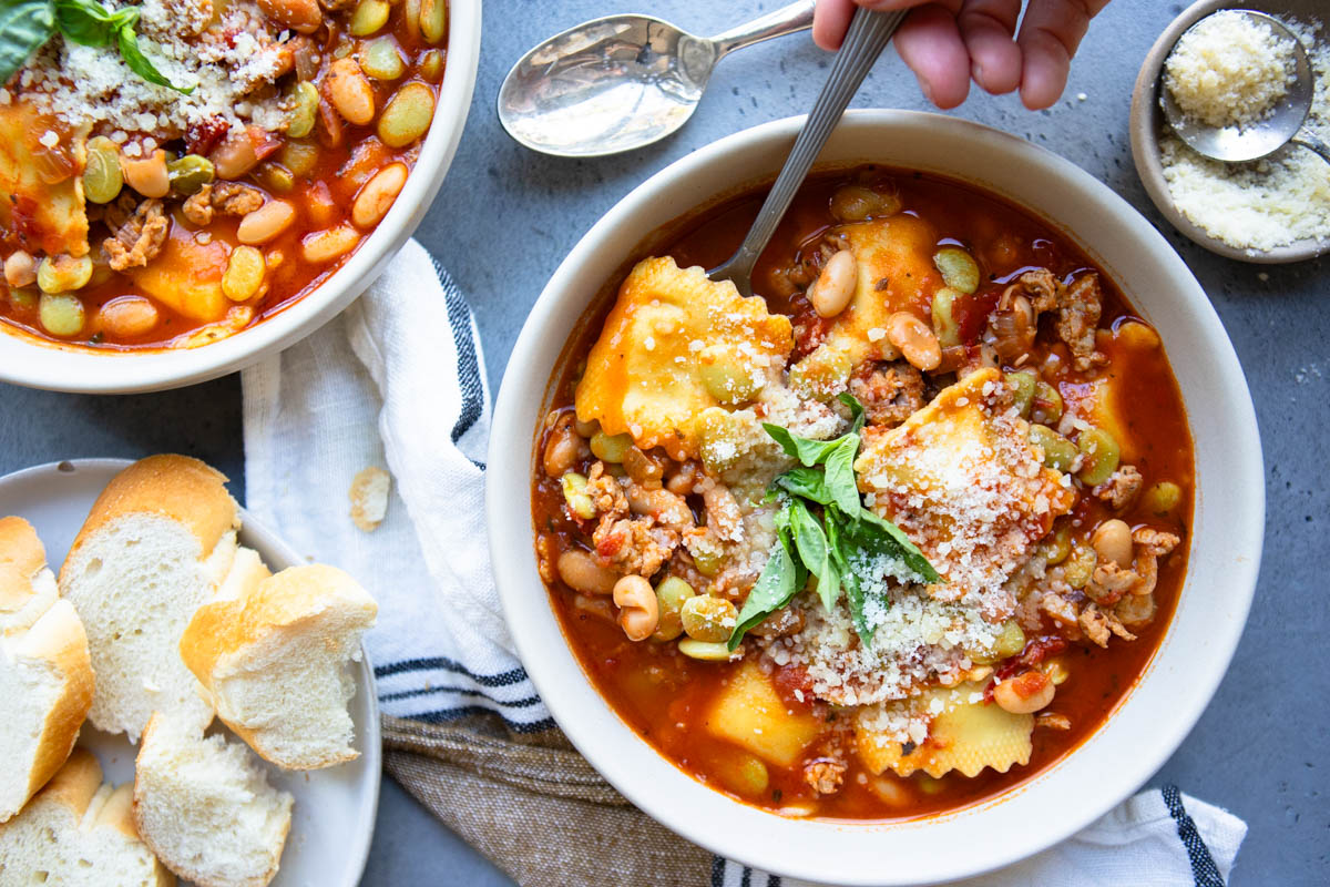 easy pasta fagioli soup recipe served in a large beige bowl and garnished with basil and parmesan cheese next to a plate of baguette bread