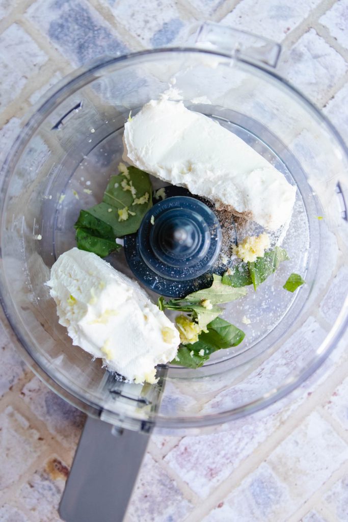cream cheese and goat cheese with garlic and basil added to the bowl of a food processor 