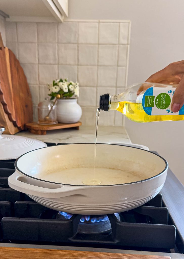 1 teaspoon canola oil being added to a white ceramic cast iron skillet