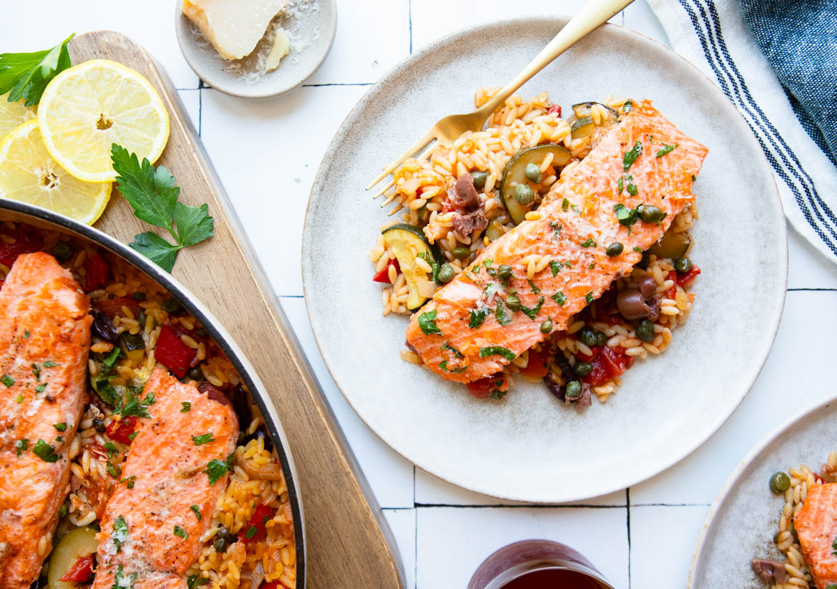 a plate filled with salmon and vegetables orzo topped with olives and capers