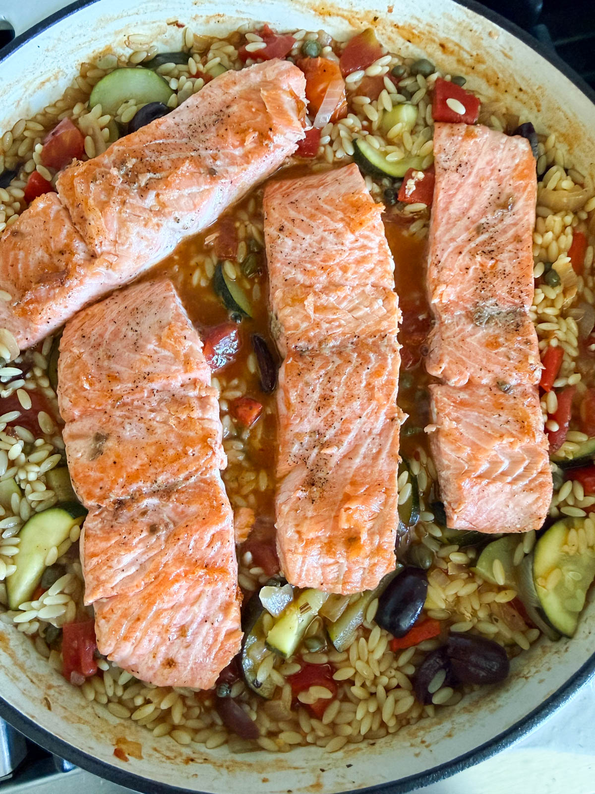 salmon filets being added to a pan filled with vegetables and orzo