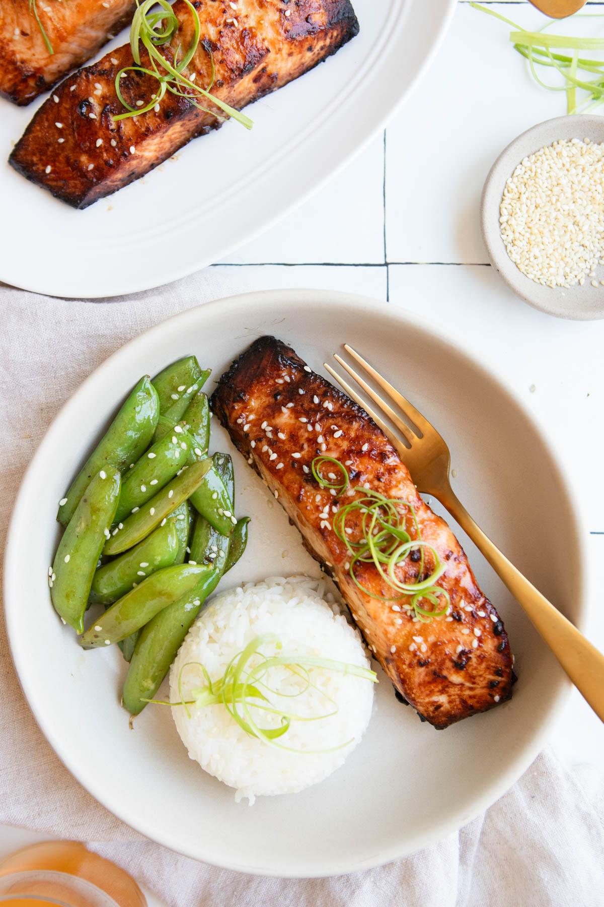 a white dish with a scoop of white rice, sugar snap peas and a piece of air fried salmon, garnished with sesame seeds.
